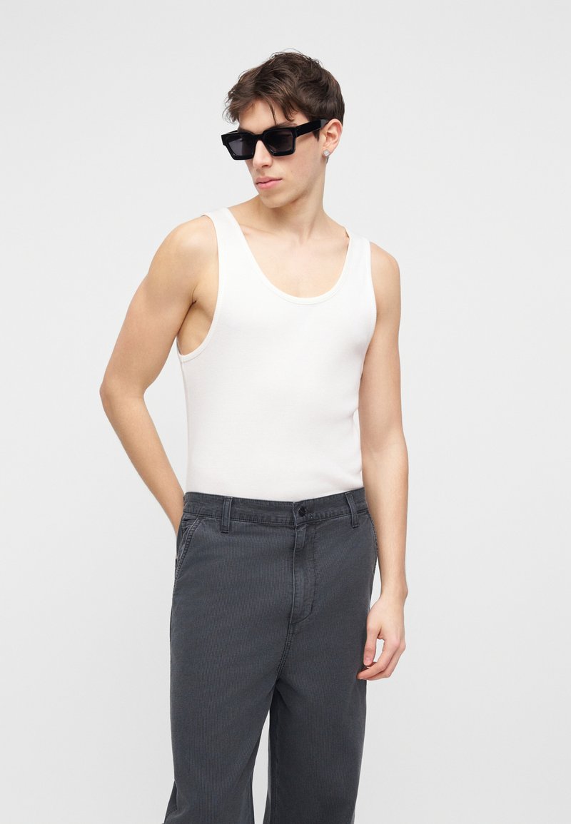 Giovane uomo con occhiali da sole neri, maglietta bianca senza maniche e pantaloni grigio scuro, in piedi con una mano in tasca su sfondo bianco.