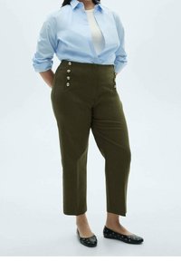 Vrouw in olijfgroene high-waisted broeken met knopen, gecombineerd met een lichtblauw shirt en zwarte platte schoenen met oogjes. 