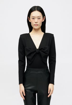 Schwarzer Bodysuit mit langen Ärmeln, tiefem V-Ausschnitt und einer großen Schleifendetail auf der Brust. Kombiniert mit glänzenden schwarzen Hosen. Glatte Textur.