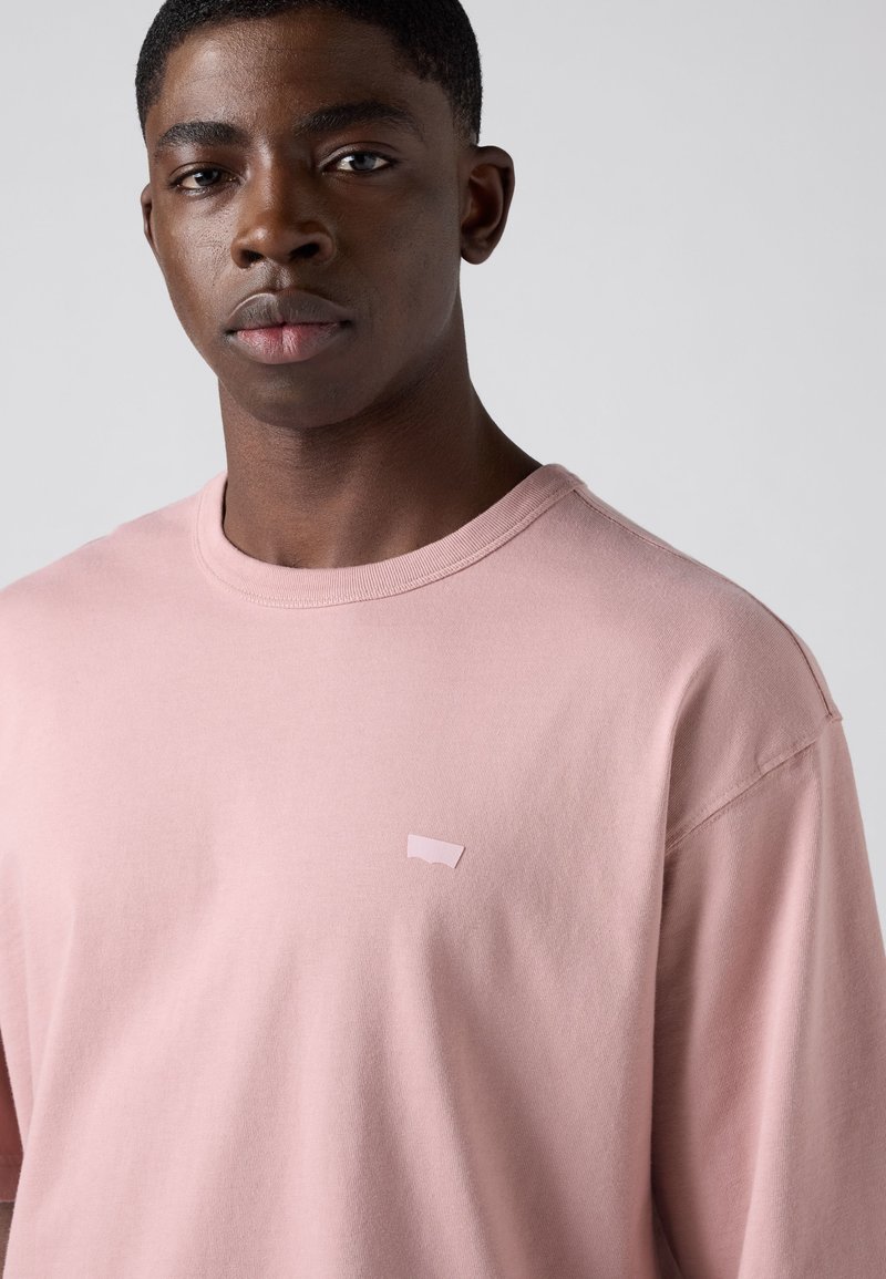 Jeune homme aux cheveux courts portant un t-shirt rose clair à col rond sur un fond uni gris clair.