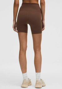 Hnedé športové šortky s vysokým pásom, hladký materiál, priliehavý strih, na dĺžku do polovice stehna, v kombinácii s béžovými teniskami a bielymi ponožkami.