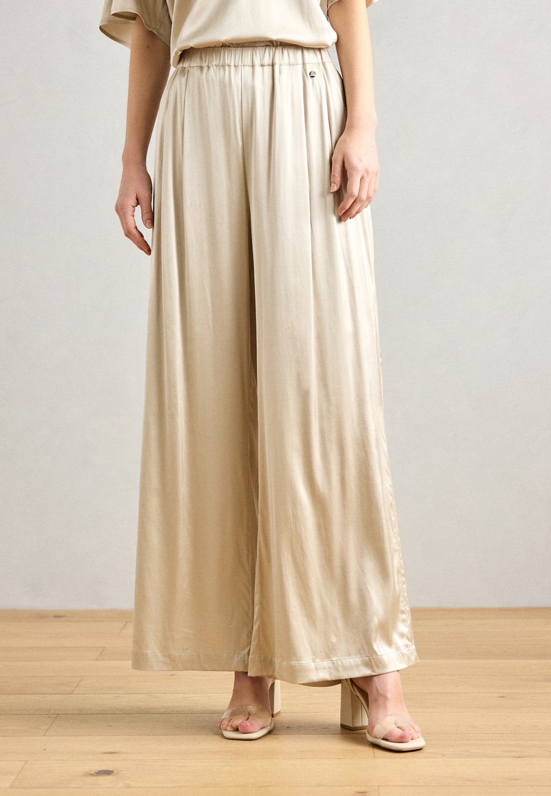 Beige wide-leg trousers made from a smooth, shiny fabric. Features an elastic waistband with a decorative button and a flowing silhouette.