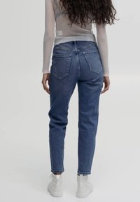Jeans de mezclilla azul claro con forma ajustada, cinco bolsillos y una cintura alta. Llevados con zapatillas blancas de caña alta sobre un fondo neutro.
