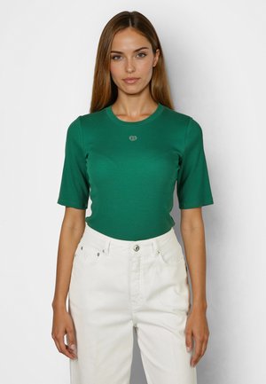 Woman with long brown hair wearing a fitted green short-sleeve shirt and high-waisted white pants against a plain background.