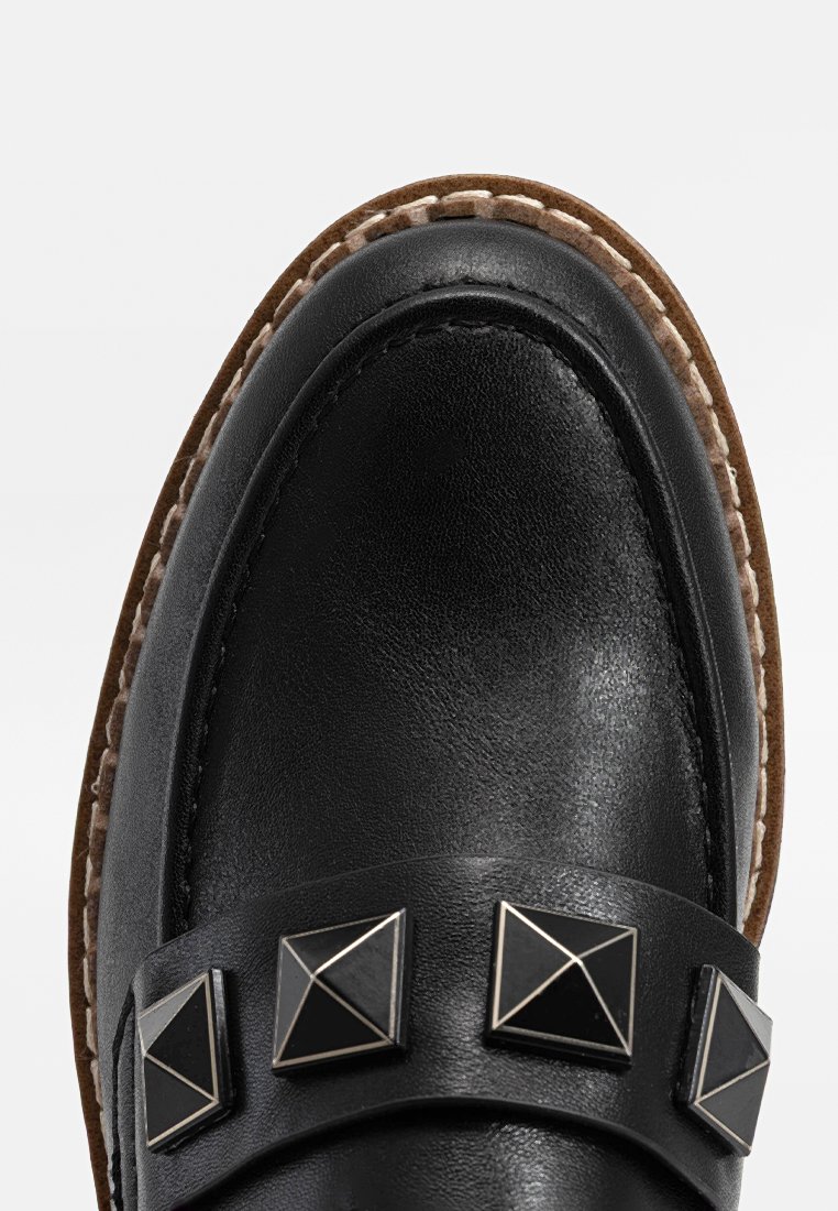 Scarpa in pelle nera con punta tondeggiante, caratterizzata da tre borchie a piramide nere su una fascia e una suola in contrasto color tan con cuciture decorative.