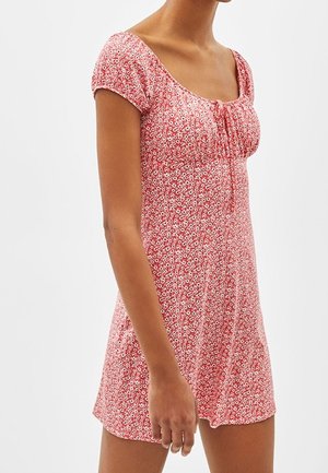 Woman wearing a short-sleeve red dress with white floral print, fitted bodice, and a scoop neckline with a small front tie.