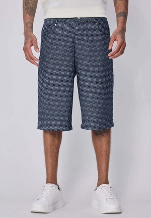 Homme portant un short bleu à motifs jusqu'aux genoux, des baskets blanches et un haut blanc, debout les bras relâchés le long du corps.