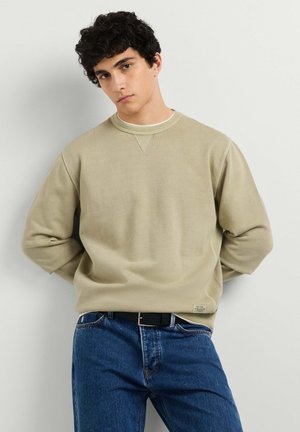 Jonge man met krullend donker haar, gekleed in een beige sweatshirt en blauwe spijkerbroek, staand tegen een effen lichtgrijze achtergrond.