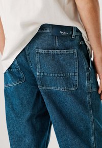 Sügavsinised teksapüksid tuhmunud viimistlusega, millel on kaks tagataskut ja vöökohal bränditud nahkmärk.