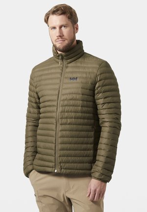 Hombre con barba que lleva una chaqueta acolchada verde oliva y pantalones beige, de pie con las manos en los bolsillos frente a un fondo liso.