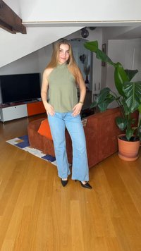 Γυναίκα με μακριά μαλλιά, φορώντας πράσινο αμάνικο τοπ και μπλε τζιν, στέκεται σε σαλόνι με καφέ καναπέ, μεγάλο φυτό και ξύλινο πάτωμα.