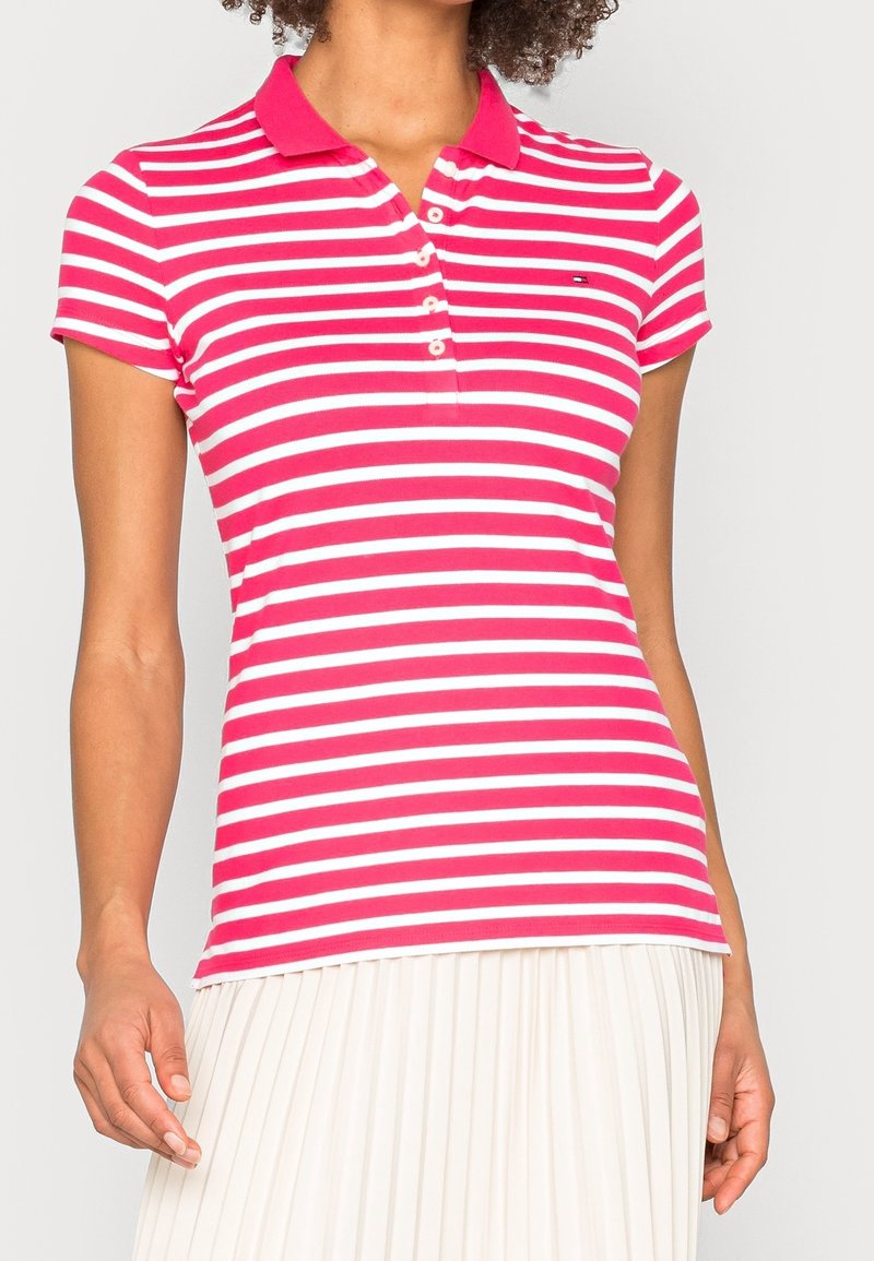Femme portant un polo ajusté à manches courtes rayé rose et blanc avec un col et quatre boutons, associé à une jupe plissée blanche.