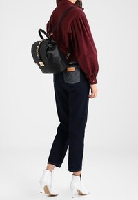 Czarny plecak ze sztucznej skóry z ozdobnym łańcuszkiem, regulowanymi paskami i złotymi elementami. Noszony z bordową bluzką i ciemnymi dżinsami.