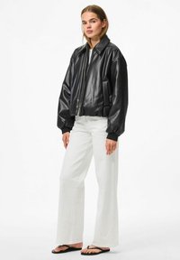 Giacca bomber in pelle nera con zip frontale, design corto, polsini a coste e tasche laterali. Abbinata a jeans bianchi a campana.