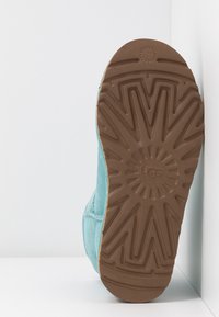 Botte en daim bleu clair appuyée contre un coin blanc, montrant une semelle en caoutchouc marron avec une bande de roulement à motif et le logo "UGG" au centre.