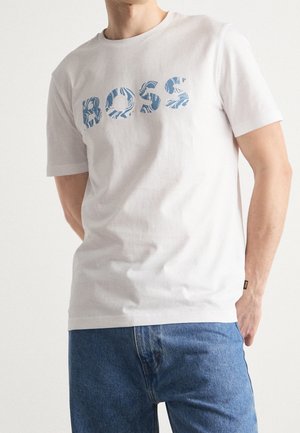 Mężczyzna w białej koszulce z niebieskim wzorzystym napisem "BOSS" i niebieskich dżinsach, stoi z jedną ręką w kieszeni na jednolitym tle.