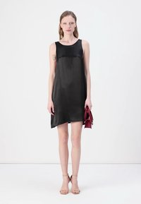 Rochie neagră fără mâneci, din satin, cu o siluetă în formă de A și un tiv asimetric. Modelul ține o mică geantă roșie.