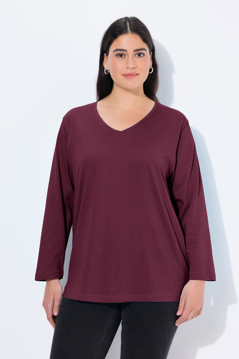 Donna con lunghi capelli scuri indossa una maglia burgundy a maniche lunghe con scollo a V e pantaloni neri, in piedi davanti a uno sfondo bianco uniforme.
