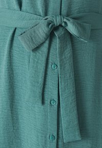 Abito in tessuto blu verde con una finitura strutturata. Presenta una patta centrale con cinque bottoni e una cintura legata in vita.