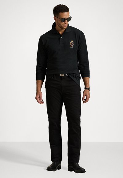 Crna polo majica dugih rukava s izvezenim logotipom, u kombinaciji s crnim trapericama i crnim čizmama do gležnja. Model koji nosi tamne sunčane naočale stoji naslonjen na neutralnu pozadinu.