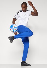 Balts sporta krekls ar melnām strīpām, zili sporta bikses ar kabatām un melni sporta apavi. Tur rokās krāsains futbola bumbas.