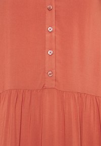 Blusa color corallo con bottoni e una texture arricciata all'orlo. Tessuto liscio con una sottile lucentezza, caratterizzata da bottoni rotondi a contrasto.