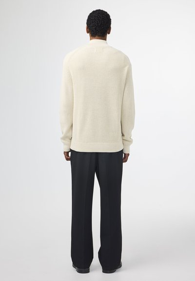 Cremefarvet strikket sweater med ribbet tekstur, der har høj krave og lange ærmer, parret med sorte vide bukser.