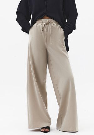 Osoba ubrana w beżowe, szerokie spodnie z elastycznym paskiem i sznurkiem, w zestawieniu z czarną bluzką z długim rękawem i czarnymi butami z odkrytymi palcami.