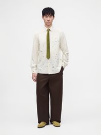 Jeune homme debout portant une chemise crème avec de petits motifs, une cravate vert olive, un pantalon large marron foncé et des chaussures jaunes sur un fond blanc uni.