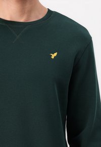 Dark green crewneck sweatshirt with small yellow bird embroidery on left chest, worn by person against a white background.