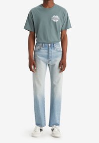 Camiseta verde de manga corta con un logo blanco, combinada con unos jeans de denim azul claro de corte recto, complementada con zapatillas blancas.