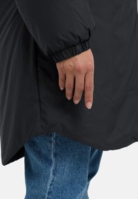 Zwarte pufferjack met elastische manchetten, zijnaaddetails en een iets langere achterkant, gedragen over blauwe denim jeans. Toont handplaatsing aan de zijkant.