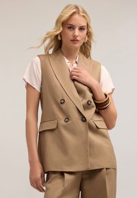 Beige kaksirivinen liivi, jossa on kapea muotoilu, sivutaskut ja mustat napit. Mallilla on päällään vaaleanpunainen paita ja rannekoruja.