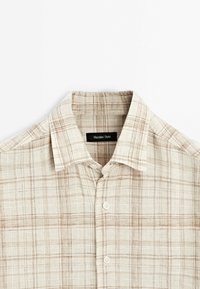 Camicia button-up a quadri chiara in beige, con motivi a reticolo marroni, colletto classico e bottoni in plastica. La trama del tessuto appare texturizzata e tessuta.