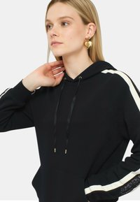 Felpa nera con cappuccio e strisce bianche sui lati, con lacci in raso e tasca frontale. Il tessuto ha una consistenza liscia.