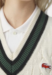 Pull en maille torsadée crème avec col en V, agrémenté d'accents à rayures vertes et noires. Logo rouge brodé en bas à gauche. Col blanc visible.