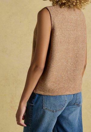 Personne aux cheveux bouclés portant un haut beige sans manches en maille et un jean en denim bleu, se tenant devant un arrière-plan beige clair.