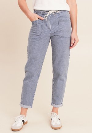 Personne portant un pantalon rayé bleu et blanc avec des poches avant et des baskets blanches, une main dans la poche, debout sur un fond clair.