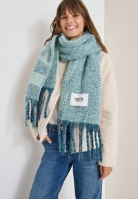 Blå strikket tørklæde med frynser, der har et struktureret mønster. Etiket læser "CECIL." Båret over en cremet farvet sweater, parret med blå jeans.