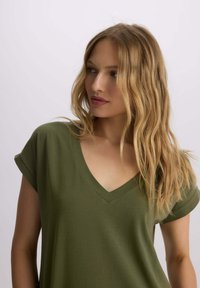 Oliwkowa koszulka typu V-neck z miękkiego materiału, z krótkimi, podwiniętymi rękawami i luźnym krojem. Gładka struktura z delikatnym wykończeniem w prążki.