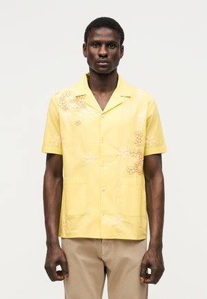 Ανδρας με σκουρόχρωμο δέρμα που φοράει κίτρινο κοντομάνικο πουκάμισο με λευκή και πορτοκαλί κεντητή διακόσμηση με λουλούδια και μπεζ παντελόνι, στέκεται μπροστά σε απλό φόντο.