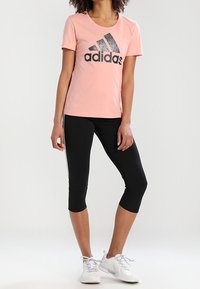 Różowy sportowy t-shirt z czarnym logo Adidas, w połączeniu z czarnymi legginsami do połowy łydki i białymi butami do biegania. Gładka tkanina, dopasowany krój.