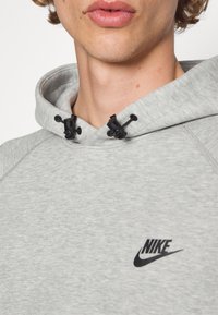 Vista parcial de una persona que lleva un hoodie gris claro de Nike con el logo negro y cordones ajustables en la capucha.