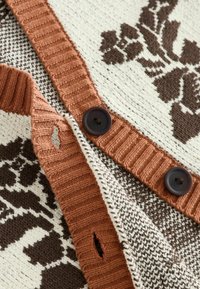 Detailní záběr hnědého a krémového pleteného kardiganu s tmavými knoflíky a květinovým vzorem na složených okrajích látky.