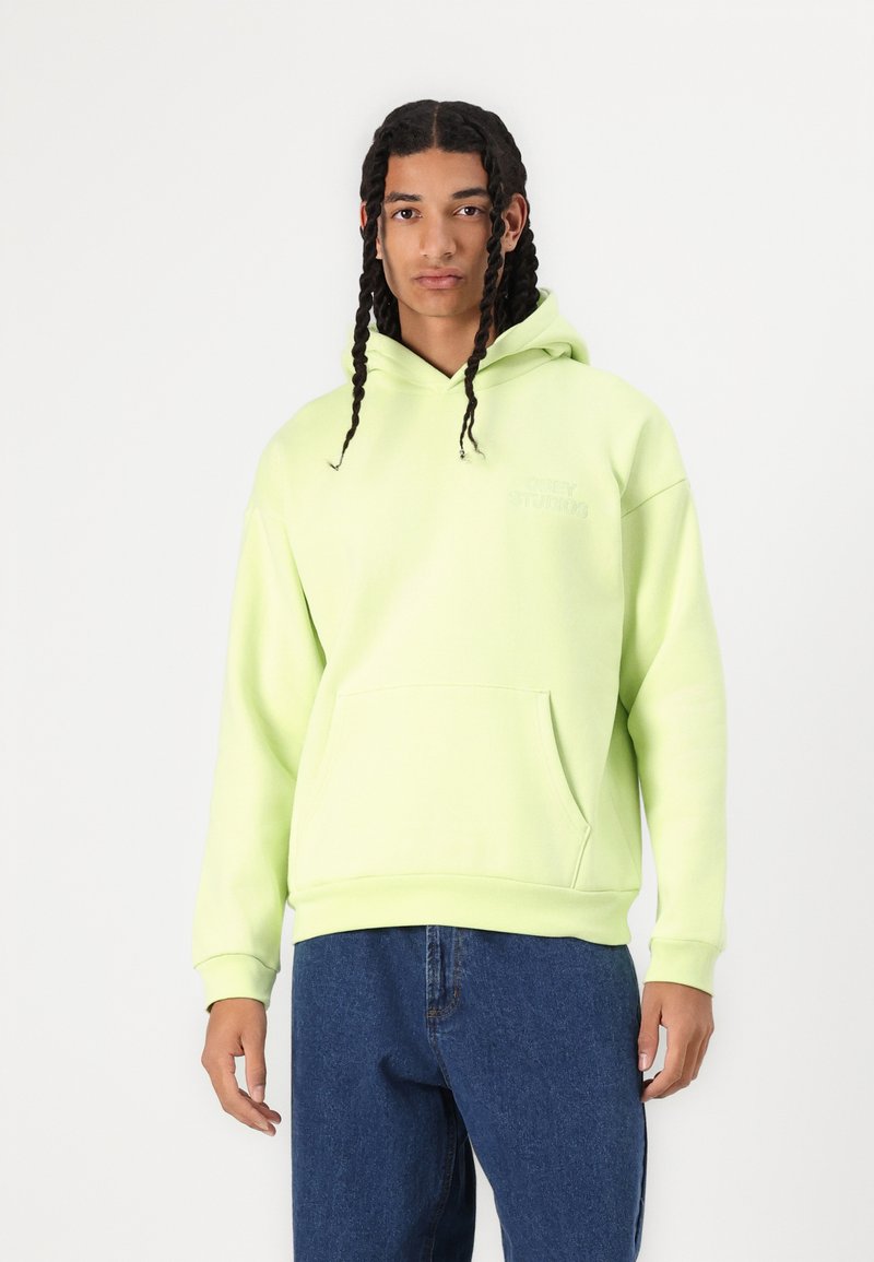 Lys grøn hoodie lavet af blødt stof, med en frontlomme, snor i hætten og broderet logo på brystet. Båret med blå jeans.