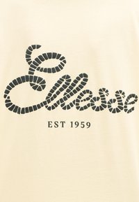 Κείμενο 'Elliot' σε μαύρο στυλ με διαχωριστικά και 'EST 1959' από κάτω σε φόντο από κρεμ ύφασμα.