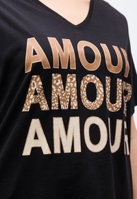 Μαύρο μπλουζάκι με V λαιμόκοψη και τη λέξη "AMOUR" επαναλαμβανόμενη τρεις φορές στο στήθος, σε μεταλλικό χρυσό, με λεοπάρ μοτίβο και σε μπεζ μπλοκ γράμματα.
