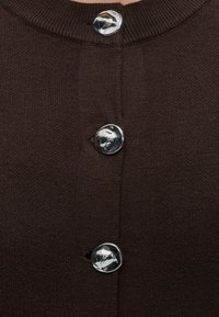 Gros plan d’un tissu tricoté brun foncé avec trois boutons argentés brillants et arrondis, fixés verticalement près du centre.