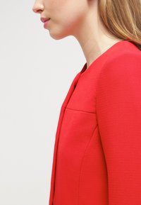 Roter taillierter Blazer aus strukturiertem Stoff, mit rundem Ausschnitt und figurnahem Schnitt. Detail einer Naht an der Schulter.