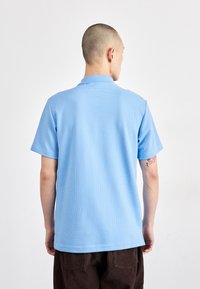 Polo de manga curta azul claro com um design texturizado e colarinho dobrado, mostrado de costas com calças de sarja castanhas.